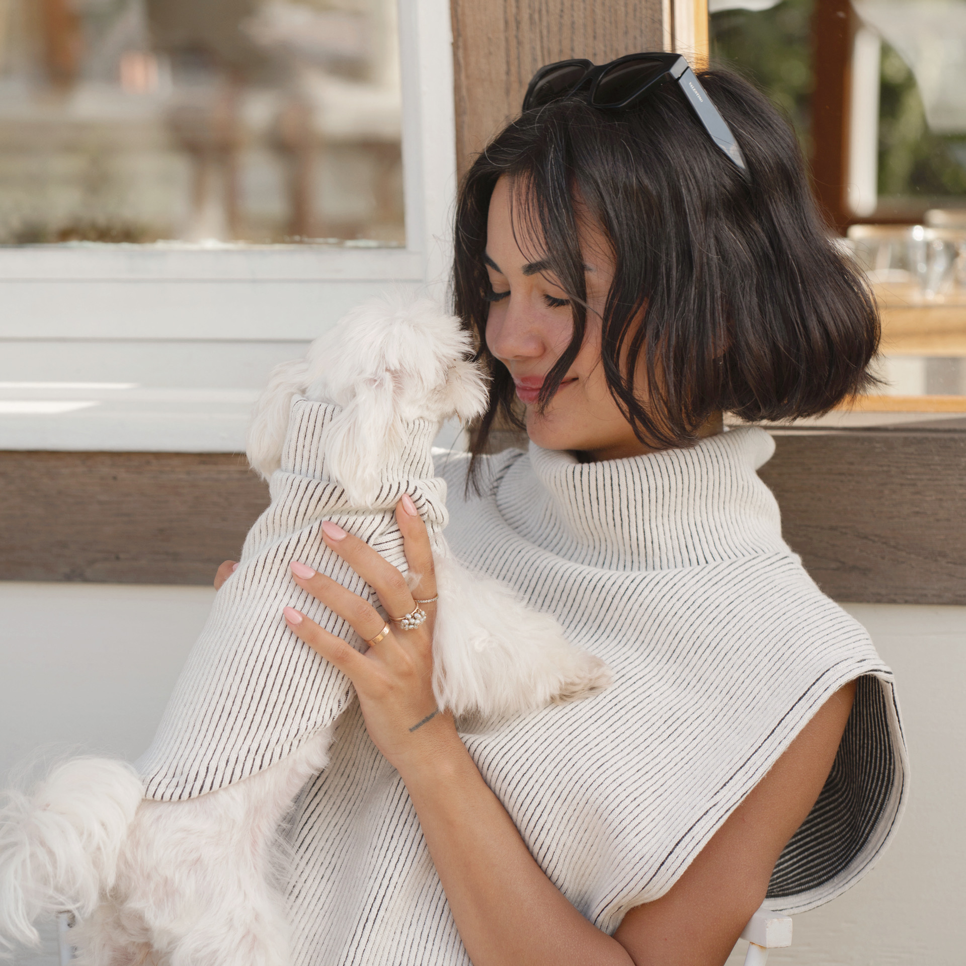 Woman sitting on a chair, holding a small white dog close to her face, wearing a cozy, sleeveless knit sweater from Lilco. The setting is warm and inviting, showcasing a moment of comfort and style.