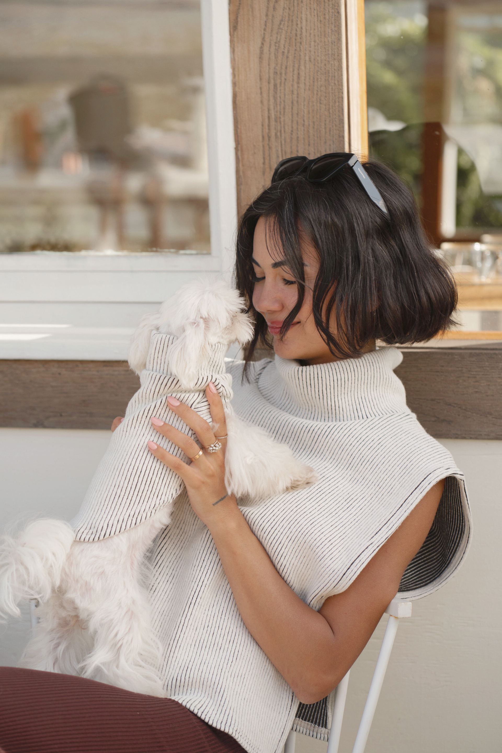 Woman sitting on a chair, holding a small white dog close to her face, wearing a cozy, sleeveless knit sweater from Lilco. The setting is warm and inviting, showcasing a moment of comfort and style.